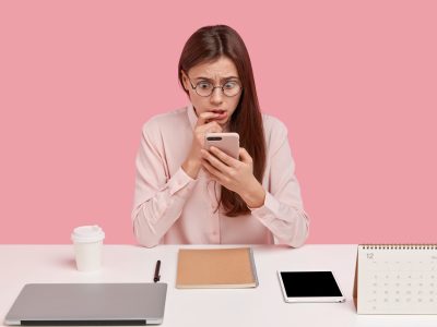 Photo of stupefied European woman browses internet on modern cell phone, has scared expression, poses at workspace, dressed in formal shirt, isolated over pink wall, reads shocking information
