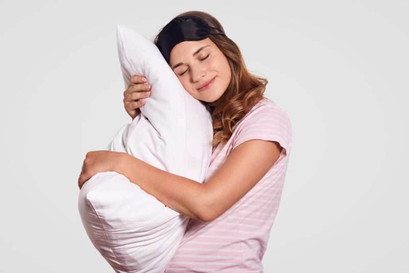 Sideways shot of good looking young woman leans on pillow, wears pyjamas and eye mask, stands against white background, has sleepy expression, wakes up early in morning. Rest and people concept